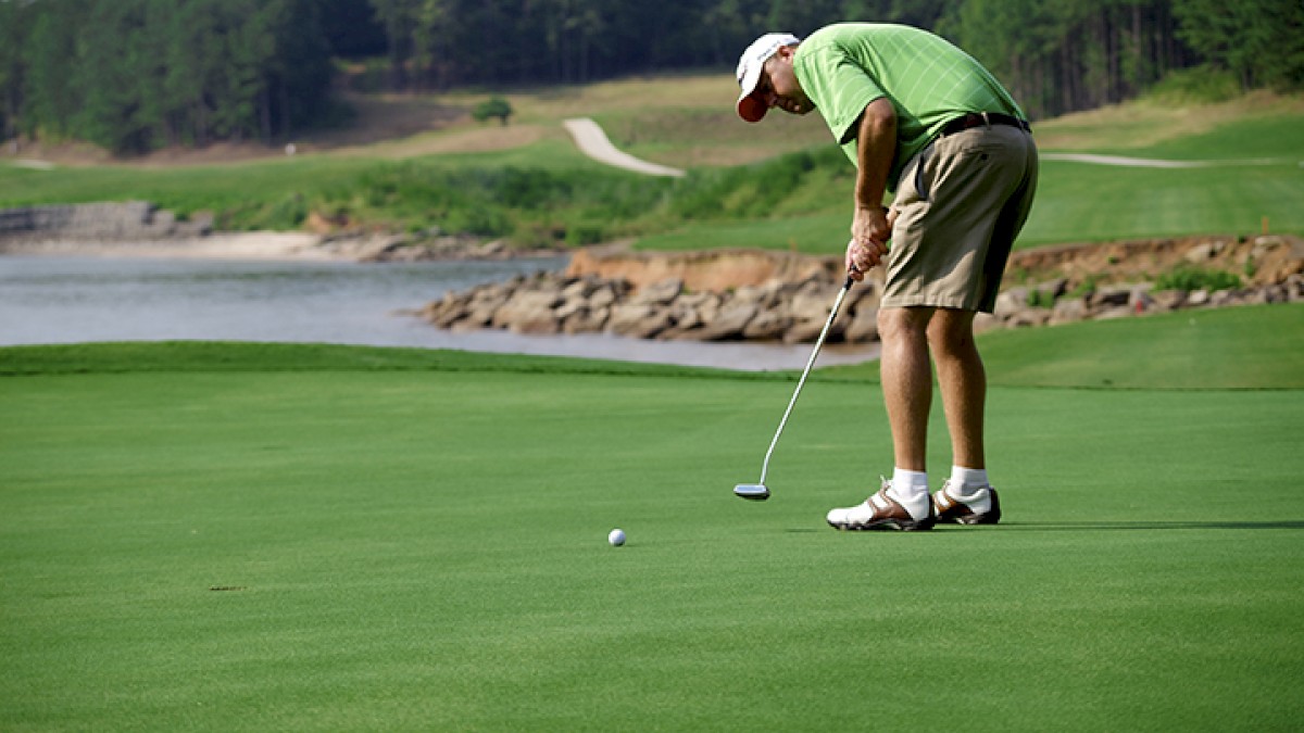 A man in a green shirt and beige shorts is preparing to putt a golf ball on a lush green golf course near water.