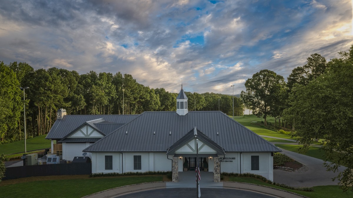 A golf club house with a black metal roof, surrounded by green trees and grass, under a partly cloudy sky during early evening.