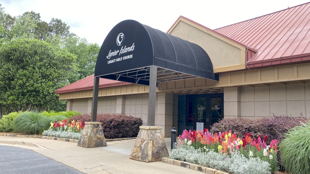 The image shows a building entrance with a black awning, surrounded by colorful flowers and greenery, and a red roof in the background.
