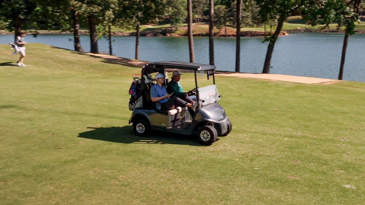 A golf cart with two individuals is on a grassy area near a lake surrounded by trees.