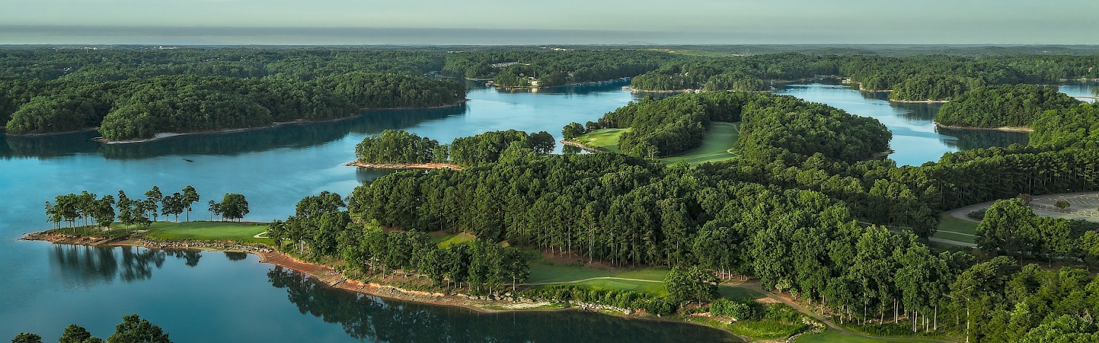 A scenic aerial view of a lush golf course surrounded by lakes and dense greenery under a partly cloudy sky.