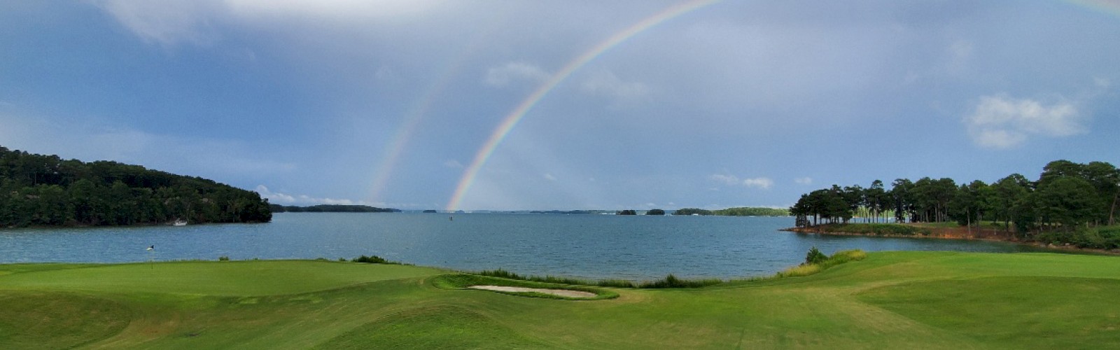 A scenic landscape with green fields, a lake, and a wide, faint rainbow arching across a cloudy sky.