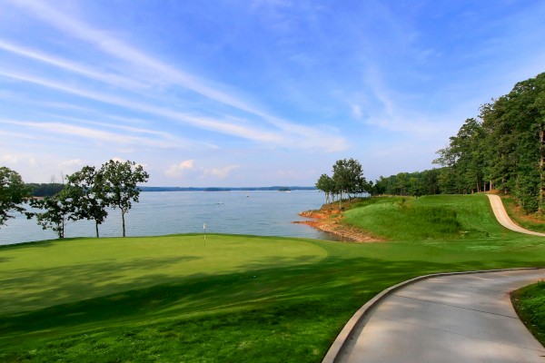 A scenic view of a golf course by a lake, with a paved path, grassy areas, trees, and a clear blue sky in the background.