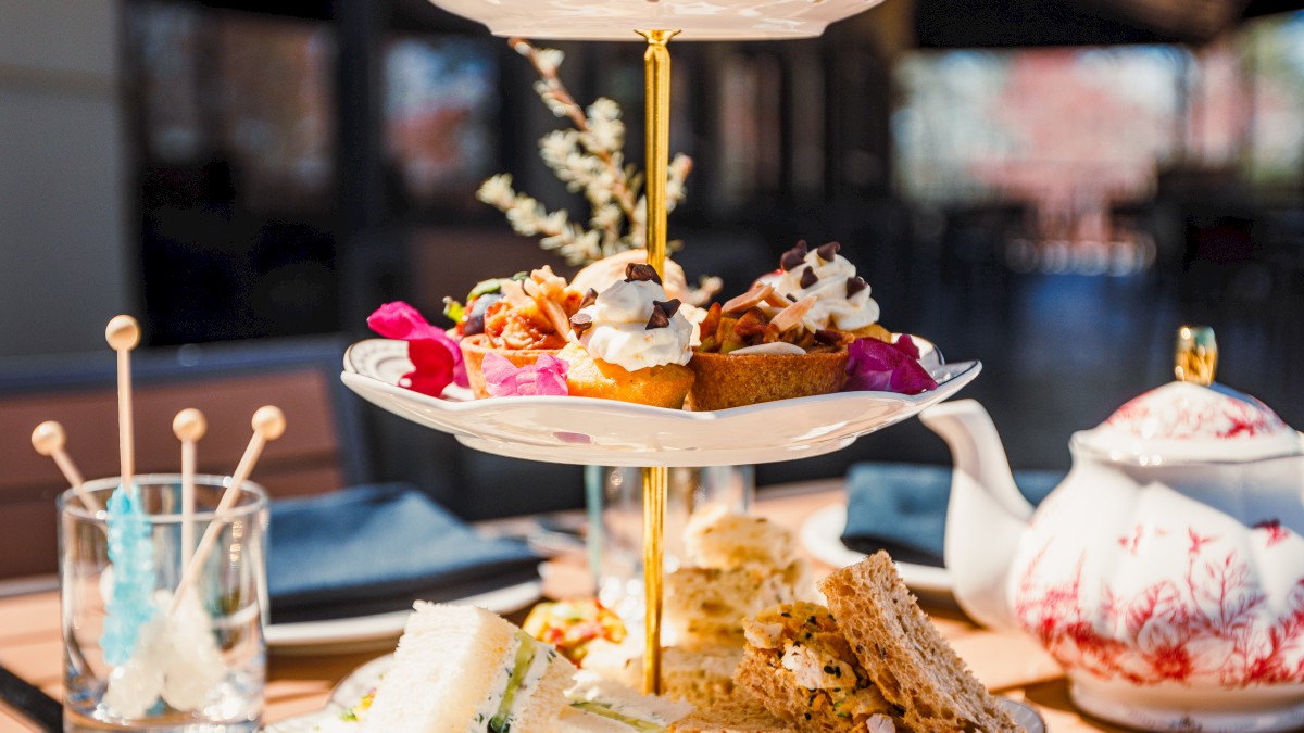 A tiered tray with assorted pastries and sandwiches is set on an outdoor table, accompanied by a floral teapot and dish.