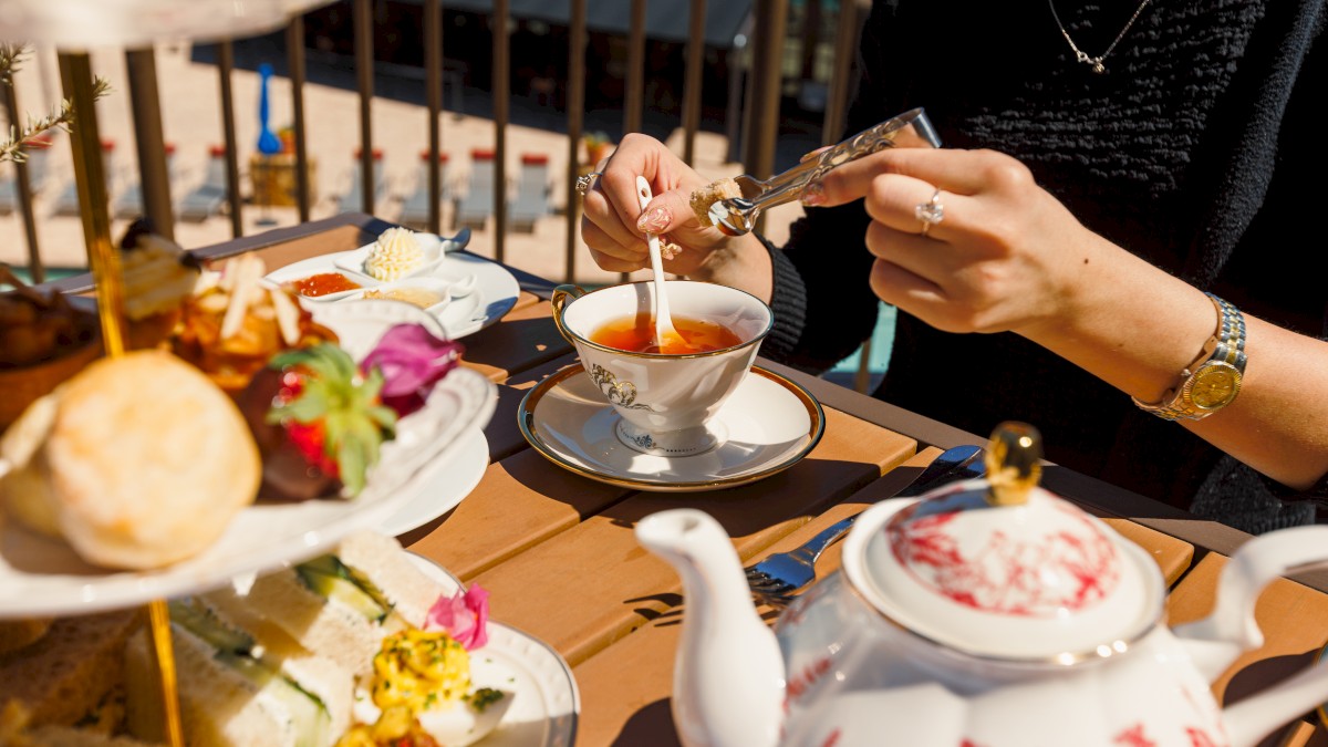 A person is stirring tea at a table with an afternoon tea setup, including a tiered tray of pastries and sandwiches, outside on a sunny day.