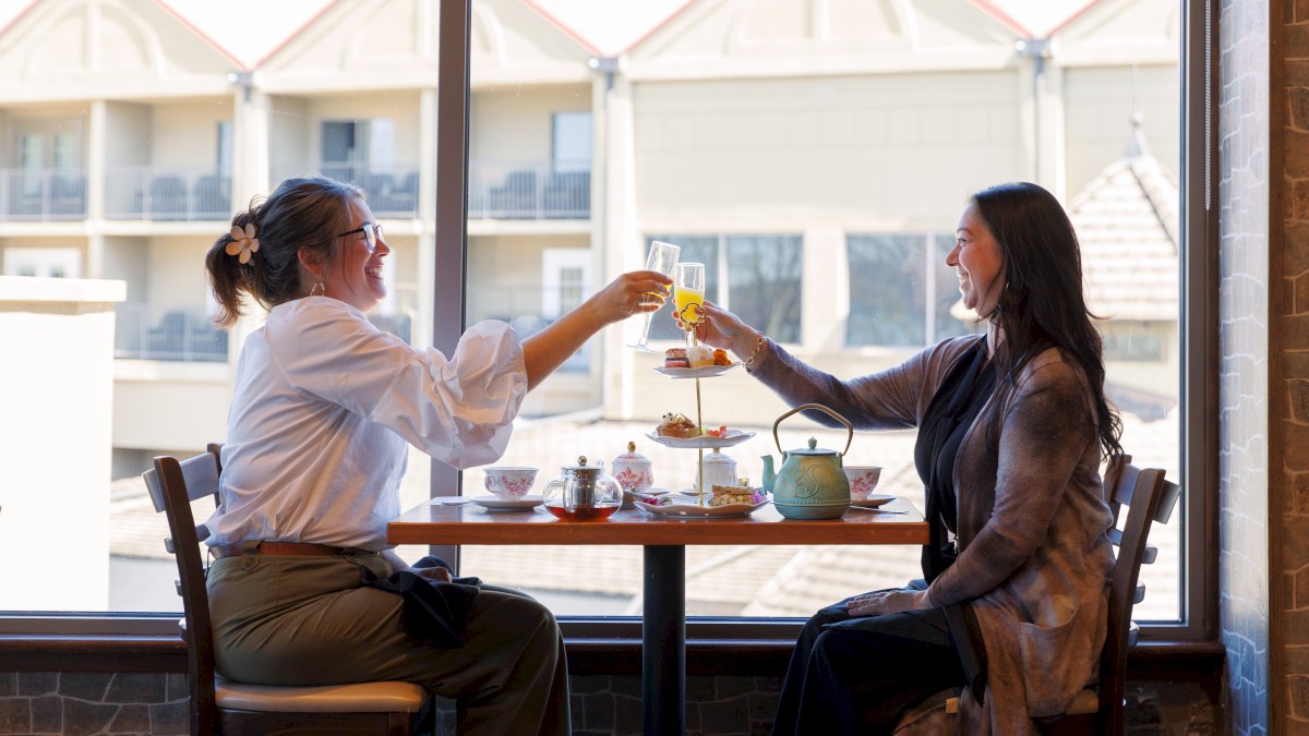 Two people are toasting at a table with drinks and a tiered tray of food, seated by a large window in a cozy setting.