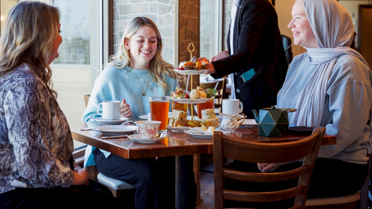 Four women enjoying tea and snacks together at a cozy cafe, being served by a lady in a blazer during afternoon.