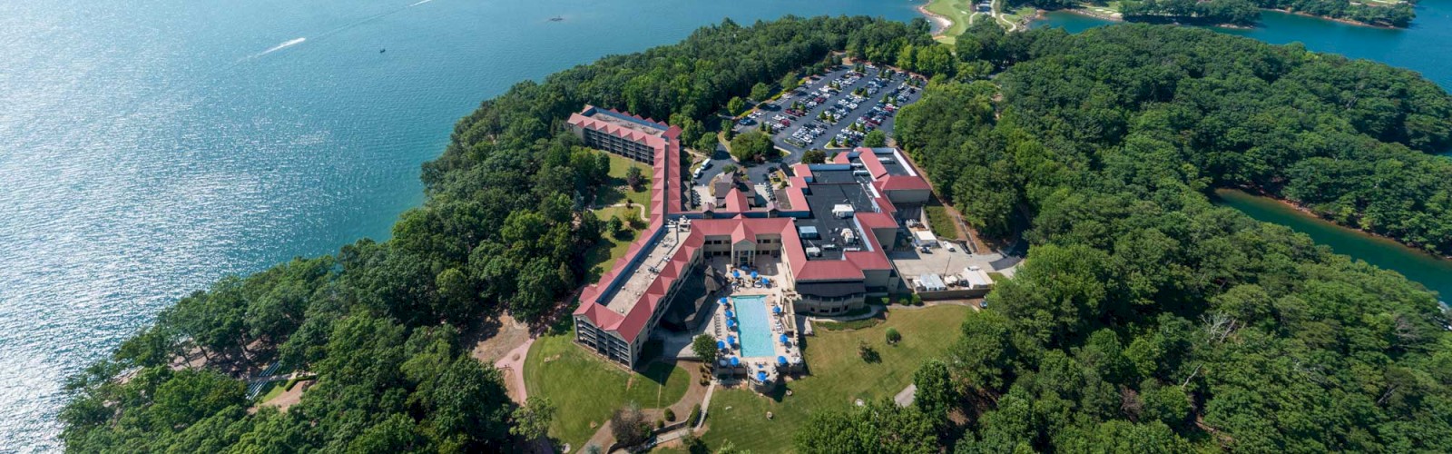 Aerial view of a lakeside resort surrounded by lush greenery, with buildings, a pool, parking area, and vast water body visible.