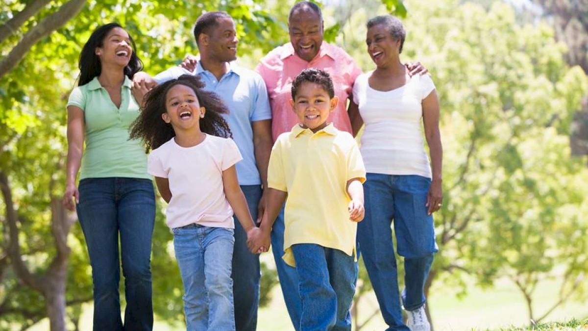 A family of six is happily walking together in a park, enjoying a sunny day with green trees in the background.