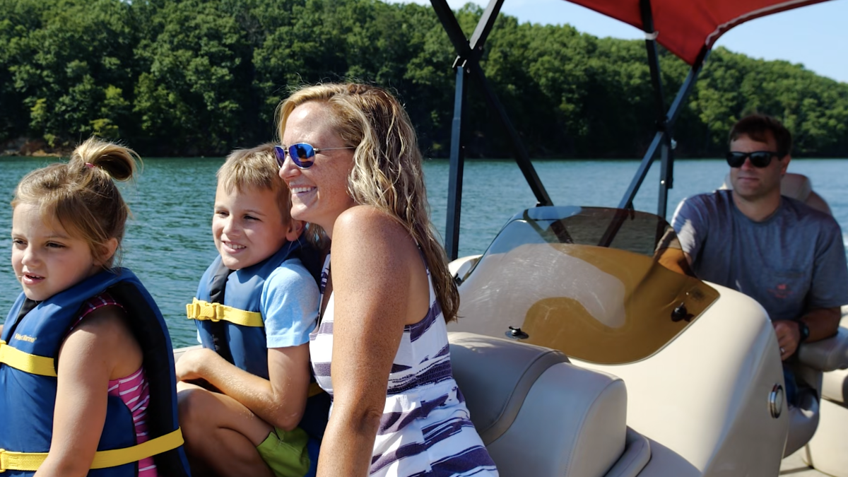 A family is enjoying a boat ride on a sunny day. The mother and two children are smiling while a man steers the boat on a lake.