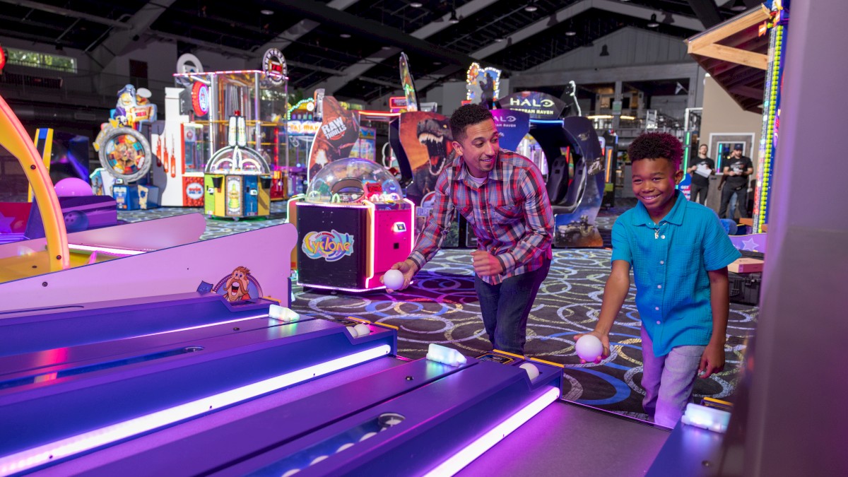 A man and a child are playing skee-ball in an arcade filled with colorful machines and games.