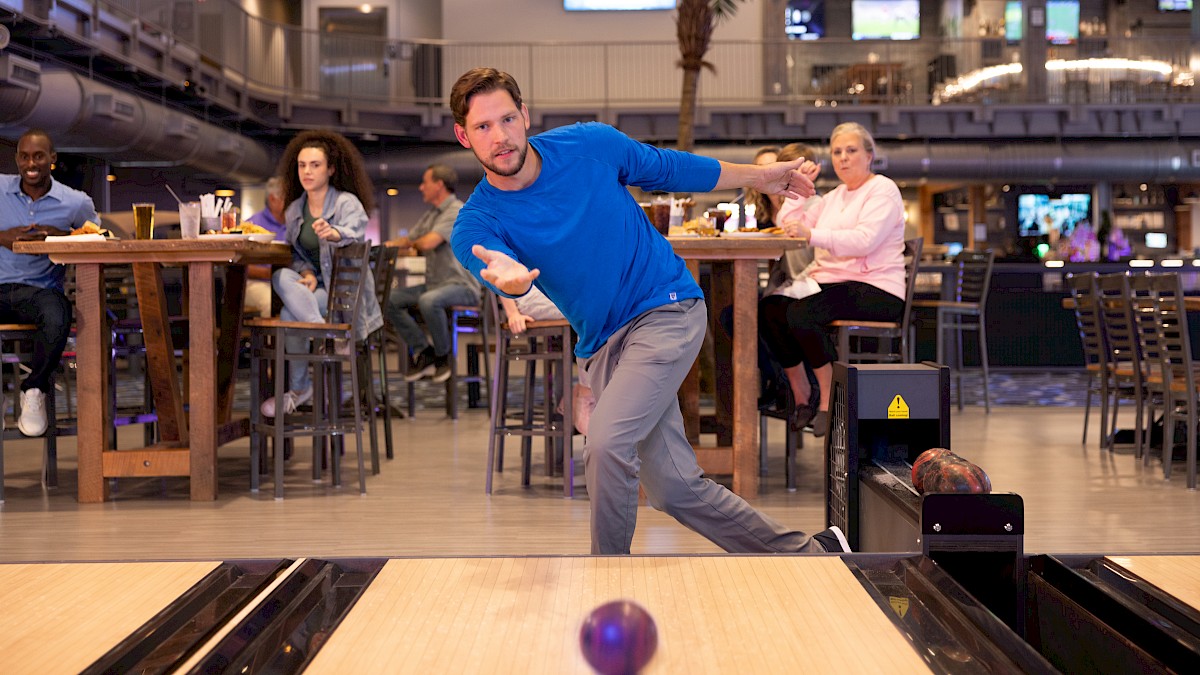 A person is bowling in an indoor venue, watched by people seated at tables nearby.