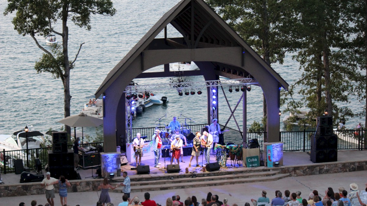 A band performing on an outdoor stage by a lake, with an audience seated in front. The setting is surrounded by trees.