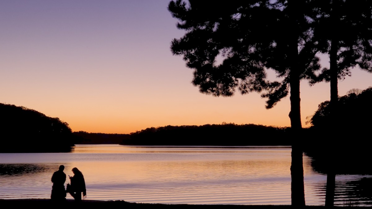 A couple's silhouette by a lake at sunset, surrounded by trees, with a colorful sky.