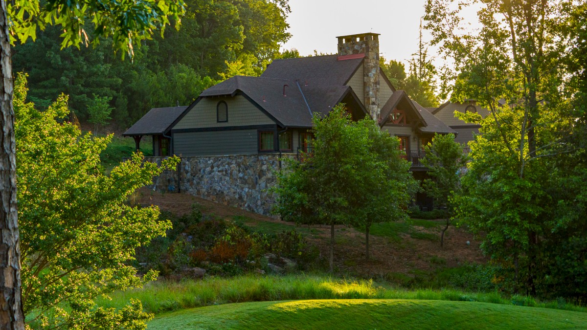 A rustic house is nestled among trees, with sunlight filtering through, surrounded by green grass and a stone wall, evoking tranquility.