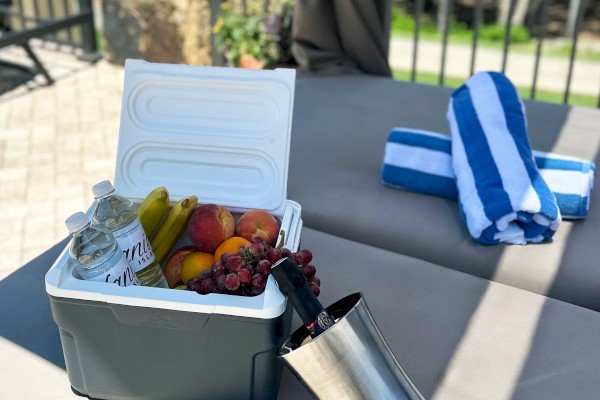 A cooler with fruit and drinks, a striped towel, and a stainless steel jug placed outdoors on a cushioned patio bench.