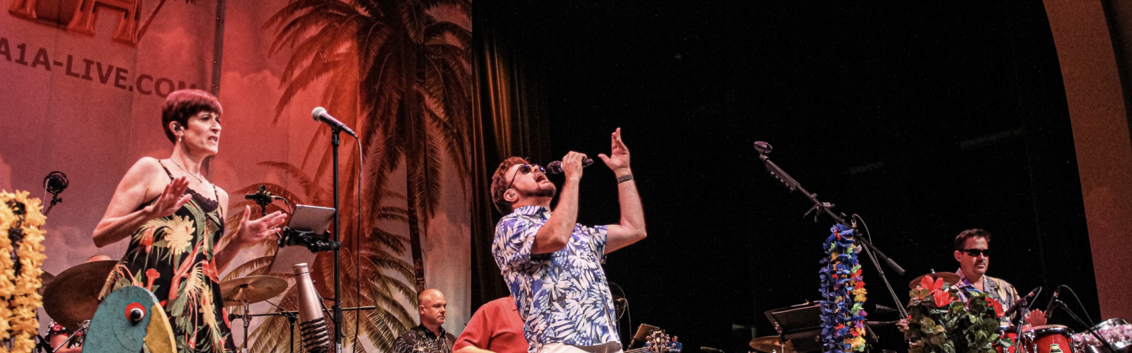 A band performs energetically on stage with tropical decorations, colorful attire, and palm trees in the backdrop.