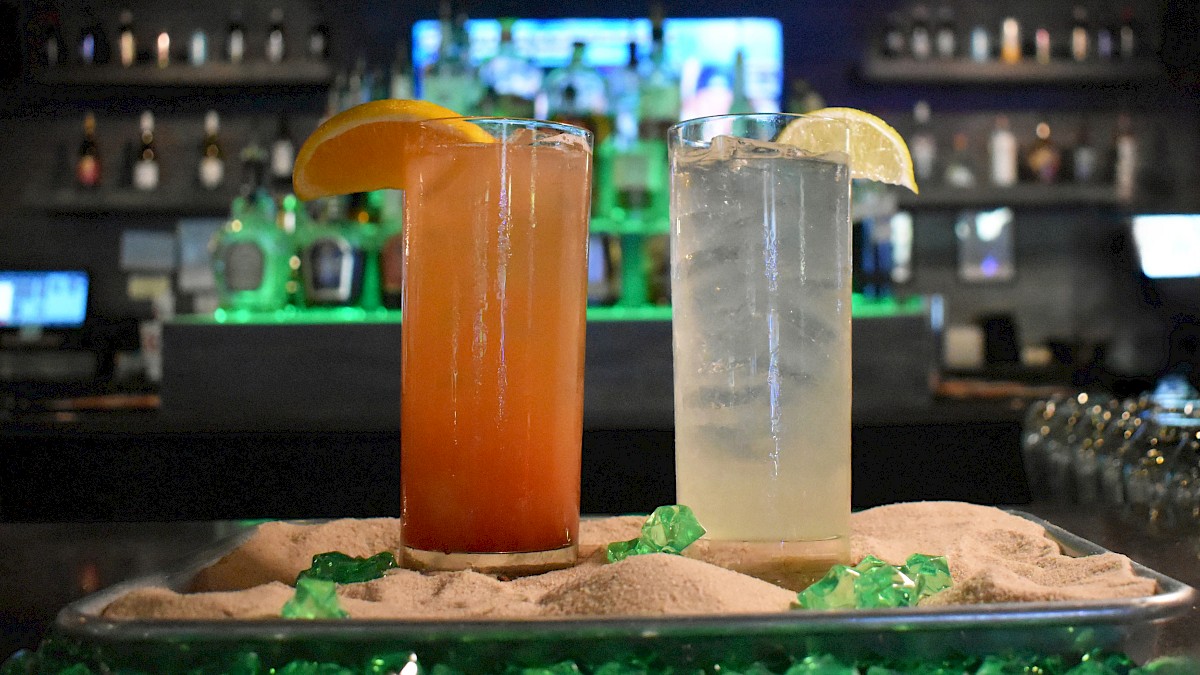 Two cocktails on a tray with sand and green decorative stones; one orange drink, one clear, each with a citrus slice garnish.
