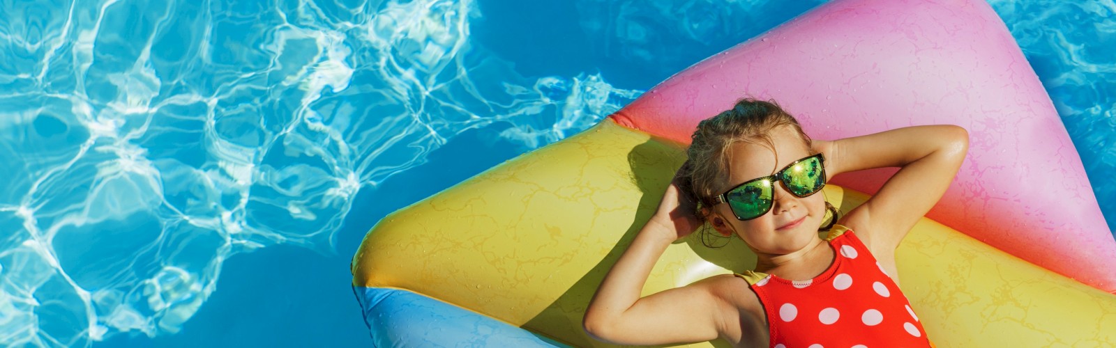A child in sunglasses relaxes on a colorful inflatable float in a pool wearing a red polka dot swimsuit, enjoying the sunny day.