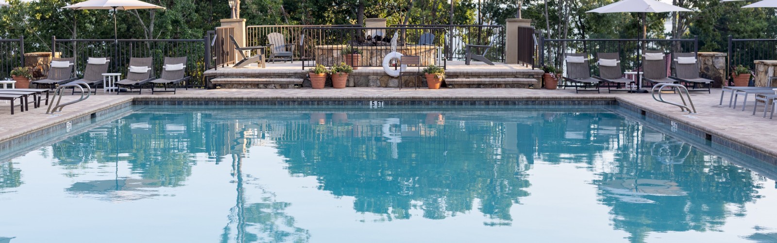 A calm outdoor pool area with clear blue water, lounge chairs, umbrellas, and trees in the background, reflecting a sunny sky.