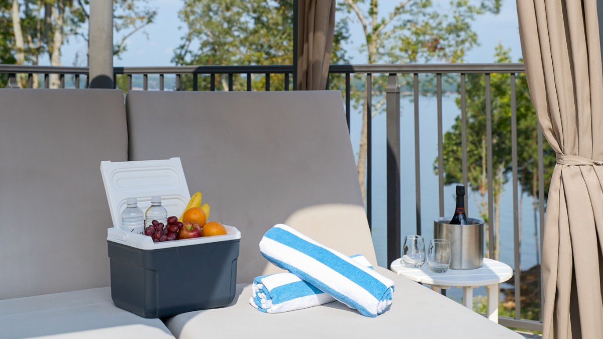 A stylish outdoor lounge on a shaded cabana with a cushioned sofa, striped towels, a small cooler with fruit, and a side table.