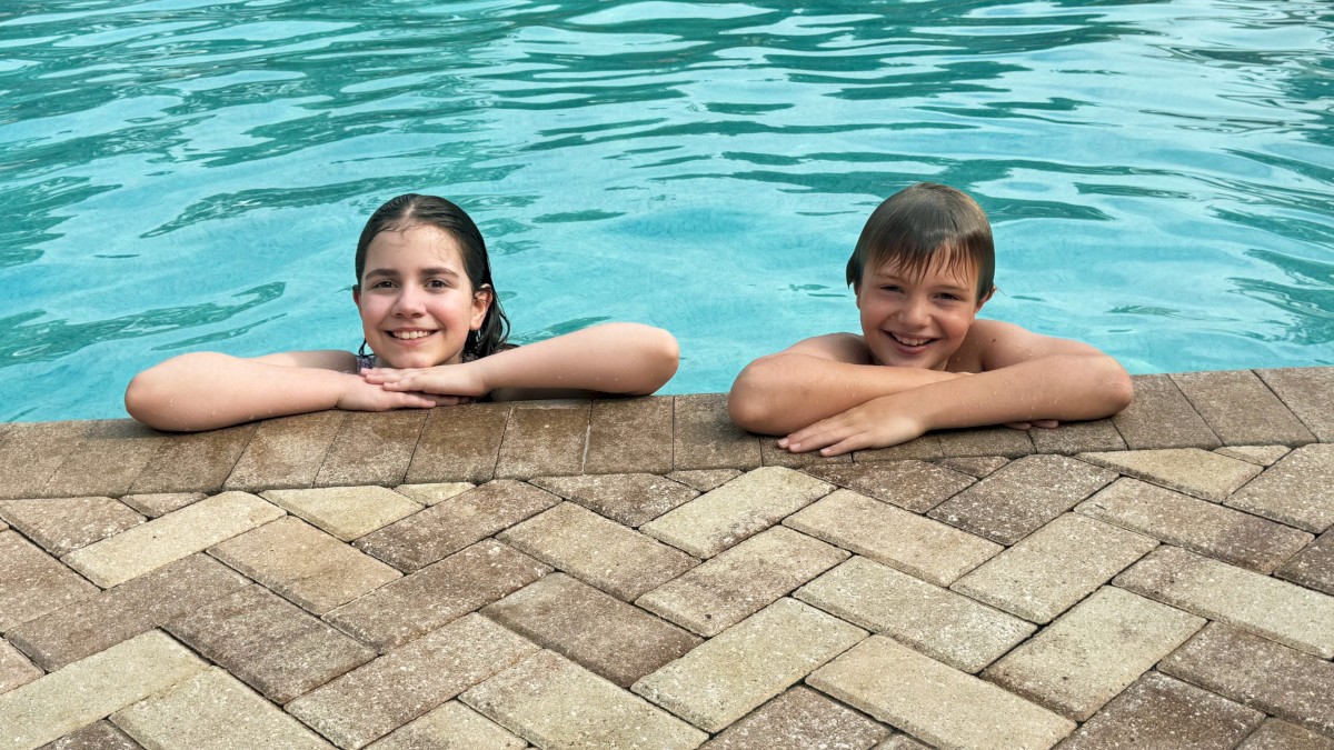 Two kids are happily leaning on the edge of a pool, surrounded by water and a brick-patterned poolside.