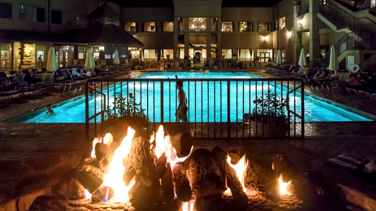 A nighttime scene with a lit fire pit in the foreground and a brightly illuminated swimming pool surrounded by loungers and a building in the background.