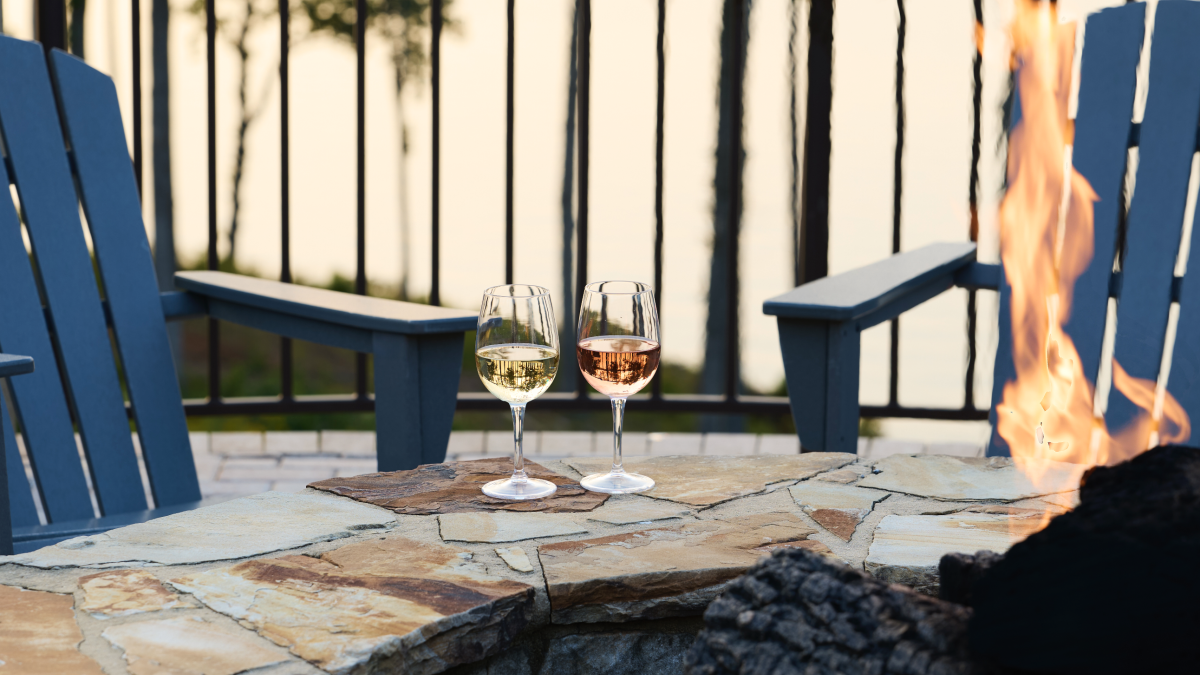 Two glasses of white and red wine on a stone table by a fire pit, with blue Adirondack chairs and a railing in the background.