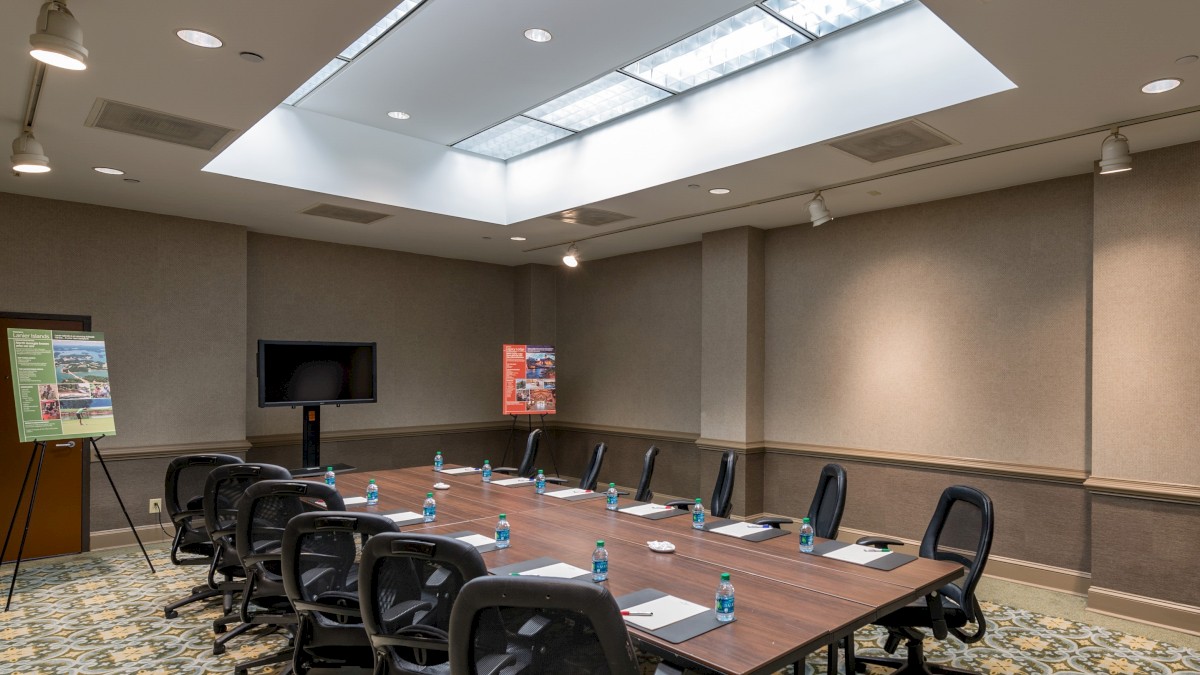 A conference room with a large table, swivel chairs, bottled water, writing pads, a TV, and presentation materials on a stand.