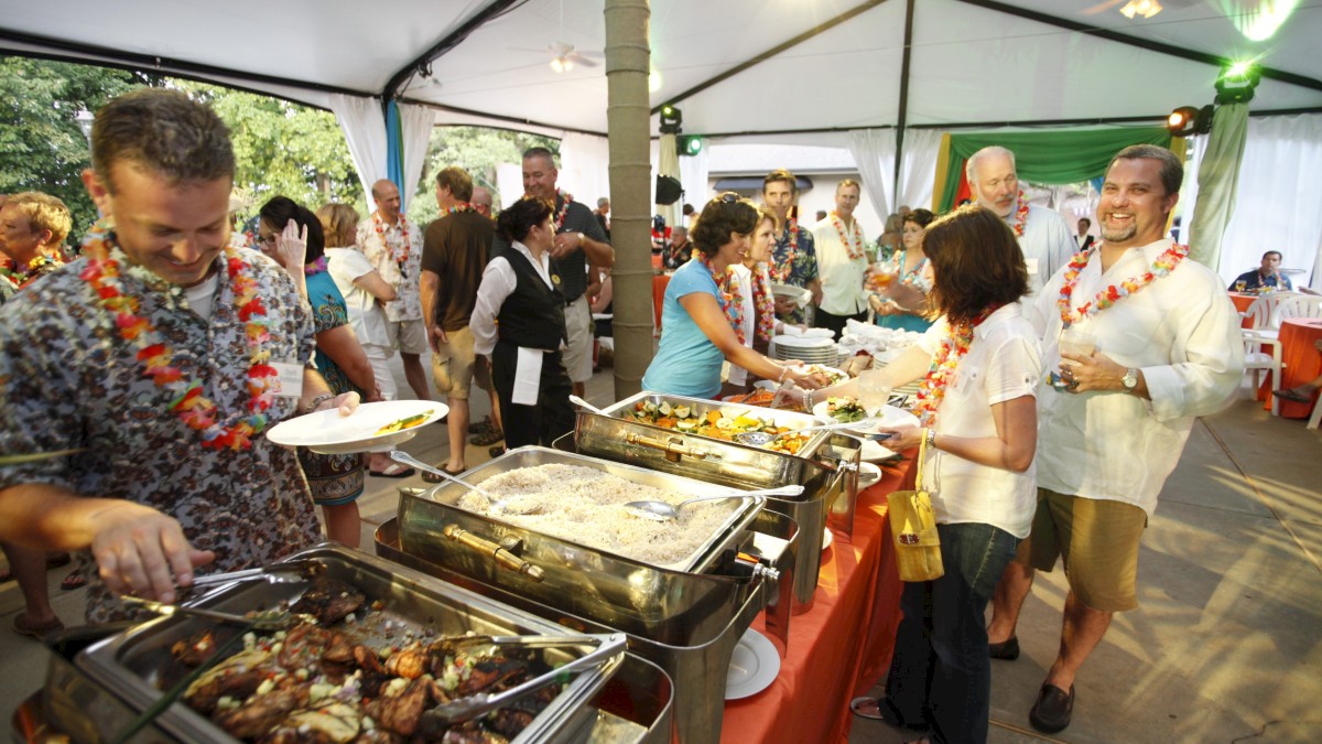 People enjoying a buffet with various dishes at an outdoor event under a tent, wearing casual attire and leis.