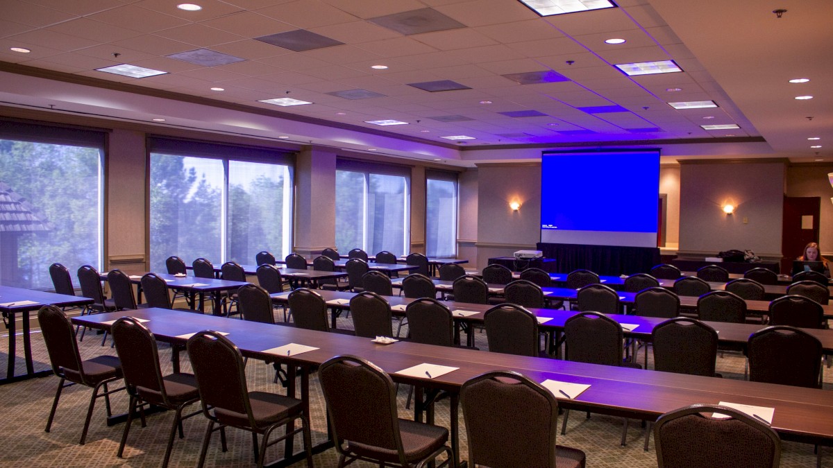 The image shows a conference room with rows of empty chairs and tables, a projection screen at the front, and large windows on the side.