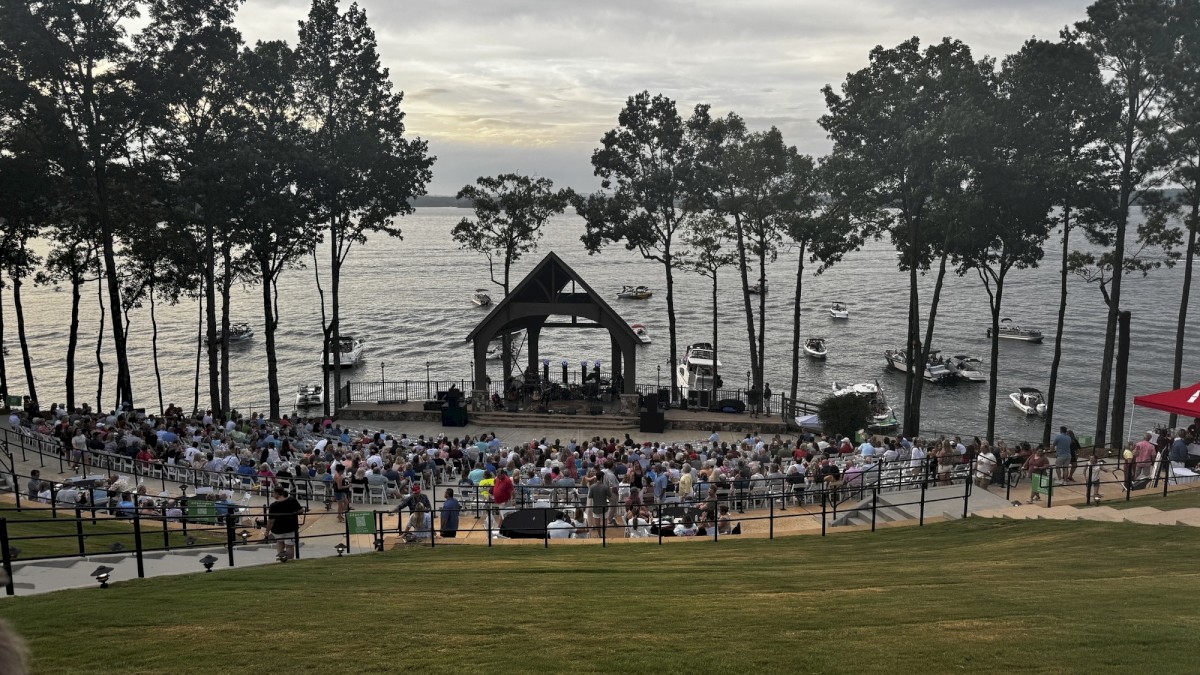 A large crowd gathers for an outdoor concert near the water, with boats floating by and trees framing the scenic lakeside setting.