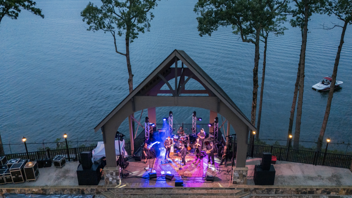 A band performs on a lakeside stage at dusk, surrounded by trees, with a boat floating on the water nearby.