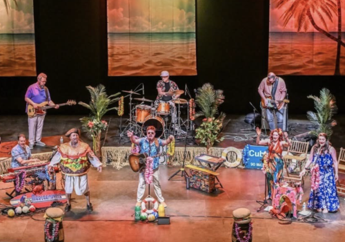 A vibrant stage performance with musicians and performers set against a tropical backdrop. Tropical decor and instruments are present.