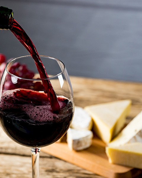 Red wine is being poured into a glass on a wooden table, with assorted cheeses and grapes in the background.