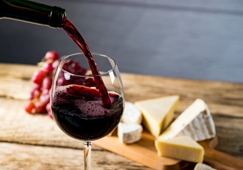 Red wine is being poured into a glass on a wooden table, with assorted cheeses and grapes in the background.