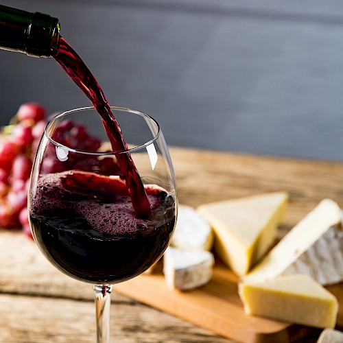 Red wine is being poured into a glass on a wooden table, with assorted cheeses and grapes in the background.