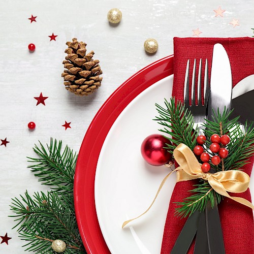 A festive table setting with a red plate, silverware wrapped in a red napkin, pine branches, berries, and decorative ornaments.