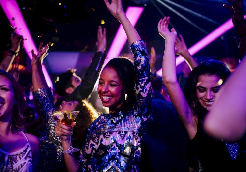 People dancing and enjoying a party, with colorful lights and confetti. Some hold drinks, smiling and raising their hands in the air.
