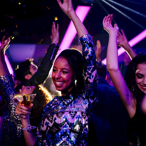 People dancing and enjoying a party, with colorful lights and confetti. Some hold drinks, smiling and raising their hands in the air.