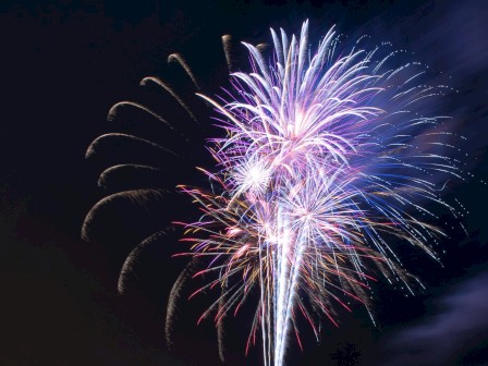A vibrant fireworks display lights up the night sky above a festive area with lit buildings and palm decorations.