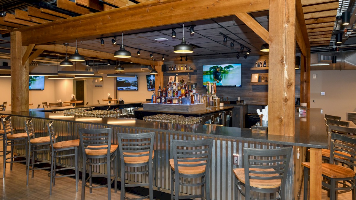 The image shows a modern bar with wooden beams, high chairs, several TVs on walls, and a variety of bottles displayed behind the counter.
