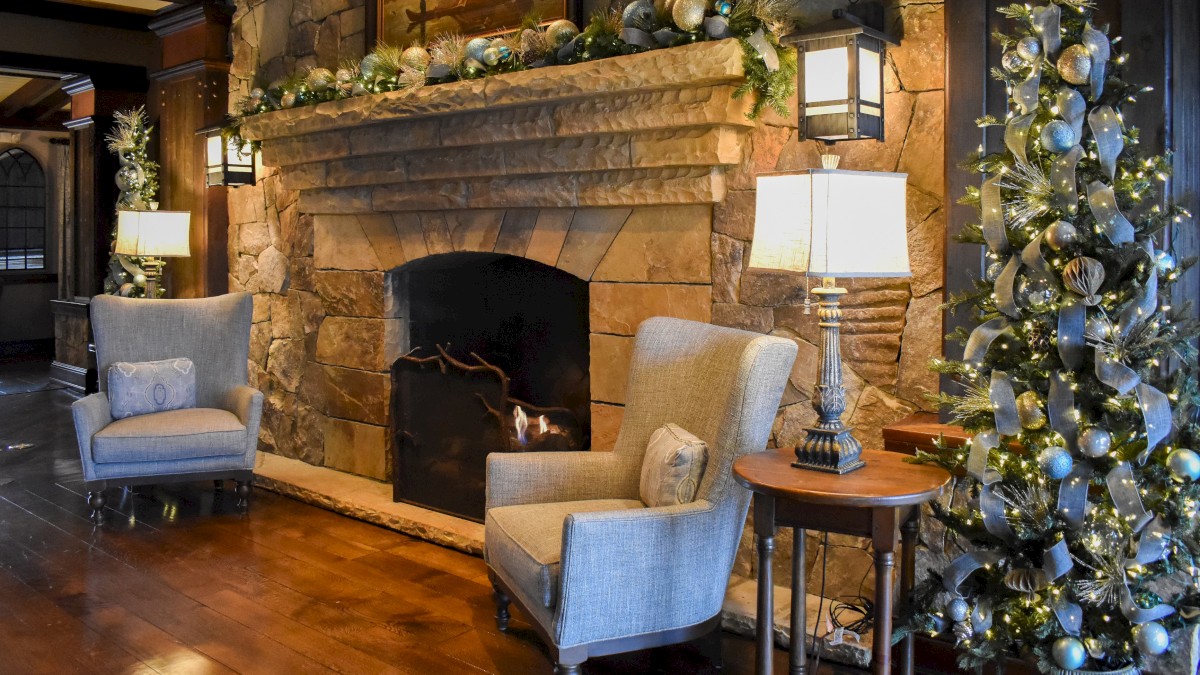A cozy living room with a stone fireplace, decorated with festive garlands, two armchairs, a side table, and a Christmas tree.