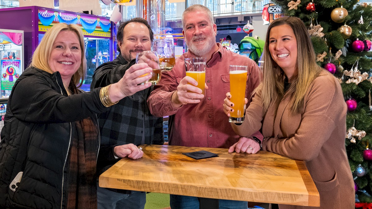 Four people are smiling and raising glasses at a wooden table in an arcade, near a decorated Christmas tree.