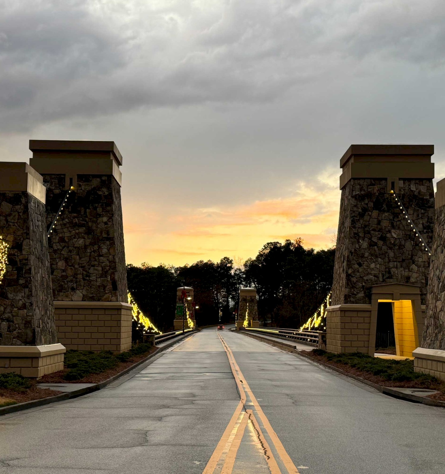 A road crosses a decorative bridge with stone pillars and lit wreaths at sunset.