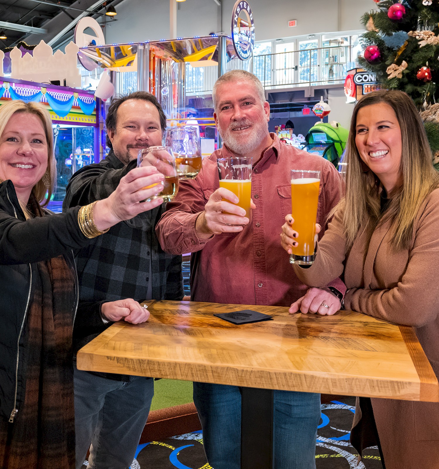 Four people are toasting with beer glasses at a wooden table. There's a Christmas tree and arcade games in the background.