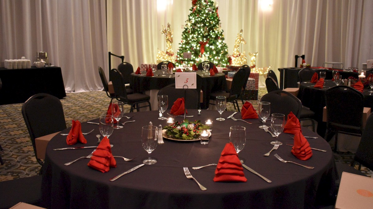 A decorated dining hall with a Christmas tree, tables set for an event, red napkins, and festive lighting completes the scene.