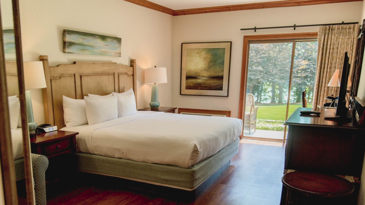 A cozy hotel room with a large bed, wooden furniture, artwork on the walls, and a sliding glass door opening to a view of greenery outside.