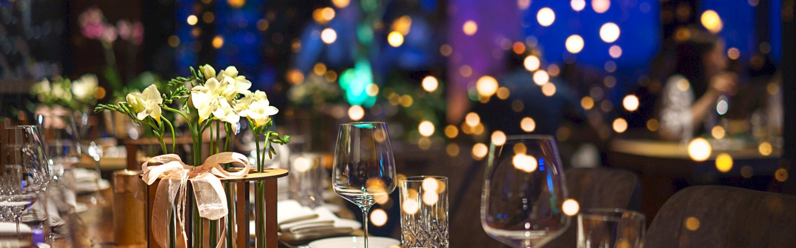 A beautifully set restaurant table with flowers, glassware, and plates, surrounded by bokeh lights creating a warm, elegant atmosphere.