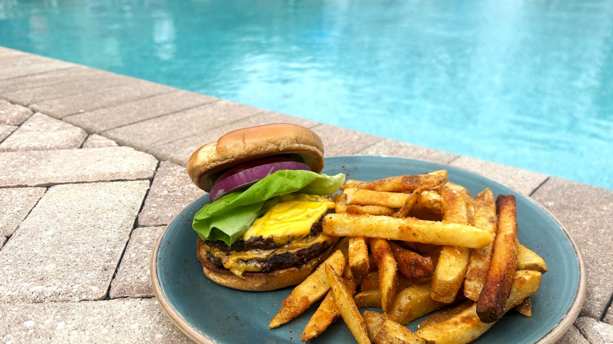 A cheeseburger with lettuce and onions, served with fries on a blue plate, placed beside a swimming pool on a tiled surface.